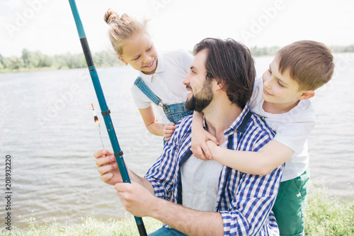 Kids are standing behind their dad, hugging and looking at him while man is holding fish-rod and looking at daughter. They are standing at river shore. They are happy.