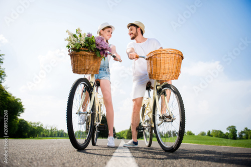 Bottom view portrait of lovely stylish couple on bikes with baskets flowers in casual outfits enjoying time together outside. Active lifestyle affection feelings daydream concept