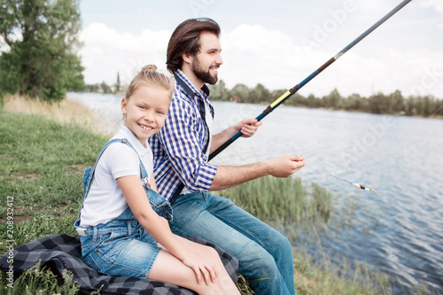 Girl is sitting besides her father and looking at camera. Guy is holding gish-rod and fishing. He is concentrated on fishing. The weather is very beautiful.