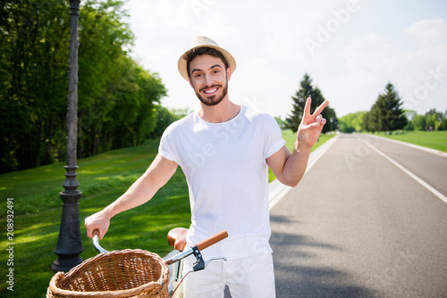 Portrait of cheerful positive attractive man in white outfit having bike gesturing two fingers v-sign with hand looking at camera. Harmony daydream serenity concept