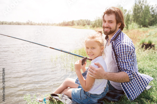 Happy dad and daughter are sitting on grass near water and looking at camera. He is huging her and holding fish-rod in right hand. They are fishing.