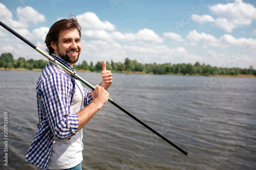 Happy guy is standing near river and looking on camera. He is smiling. Guy is holding fish-rod with right hand on right shoulder. Also man is pointing up. He looks happy.