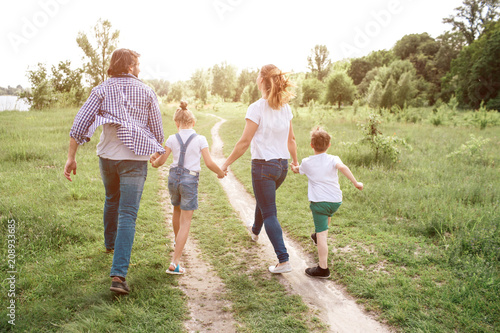 Happy family is walking down the road in meadow. Parents are holding their kids by hands. They are jumping and enjoying the moment.