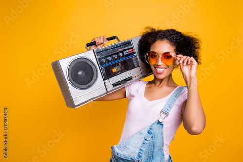 Canvas Print Portrait of fancy toothy girl with beaming smile in eyewear holding boom box on shoulder looking at camera isolated on yellow background