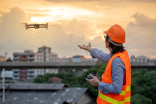 Young Asian engineer flying drone over construction site during sunset. Using unmanned aerial vehicle (UAV) for land and building site survey in civil engineering project.