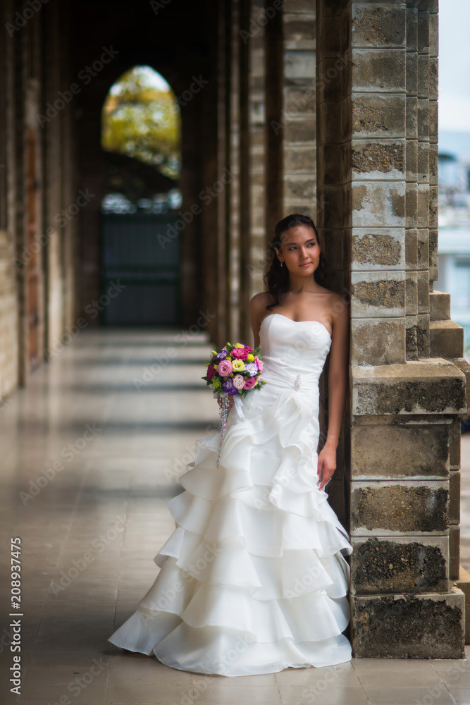 The bride stands in a gallery of stone walls in a beautiful white wedding dress with a bouquet of flowers and looks away
