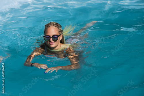 A young girl in a blue swimsuit in sunglasses sexually posing in the pool  in clear blue water-the concept of lifestyle
