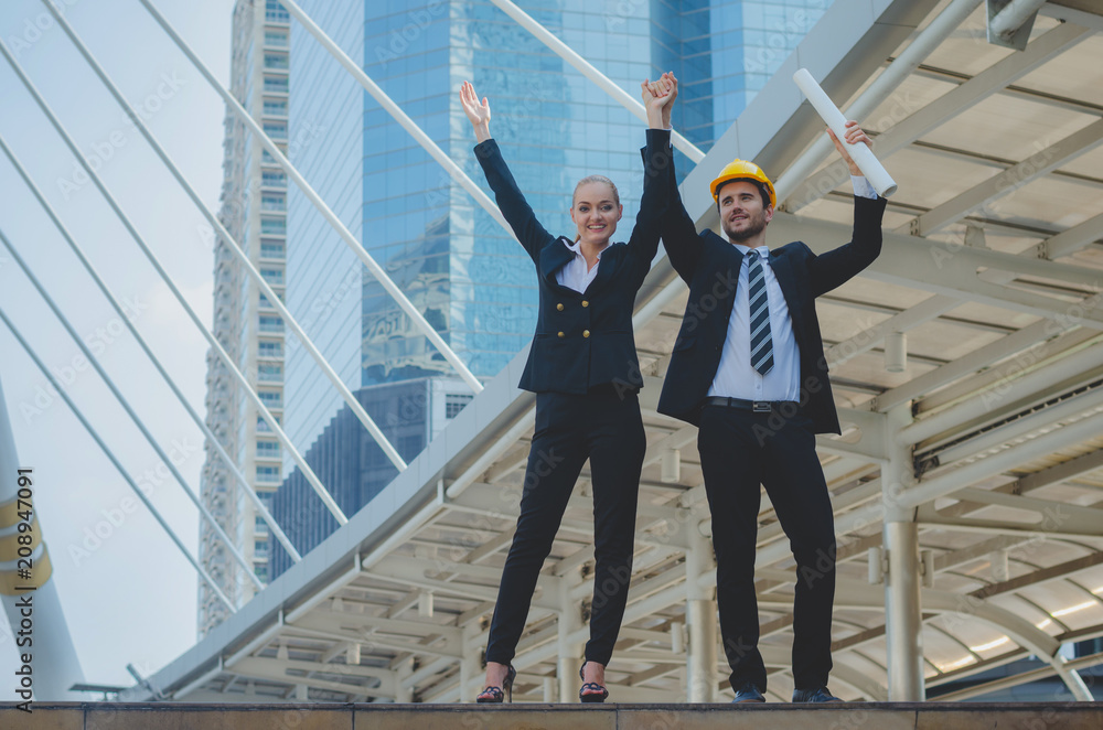 engineer man and businesswoman arms up with smile and happy together for winner and successful at outdoor with building background in city. business successful winner together concept. engineer man and businesswoman arms up with smile and happy together for winner and successful at outdoor with building background in city. business successful winner together concept.