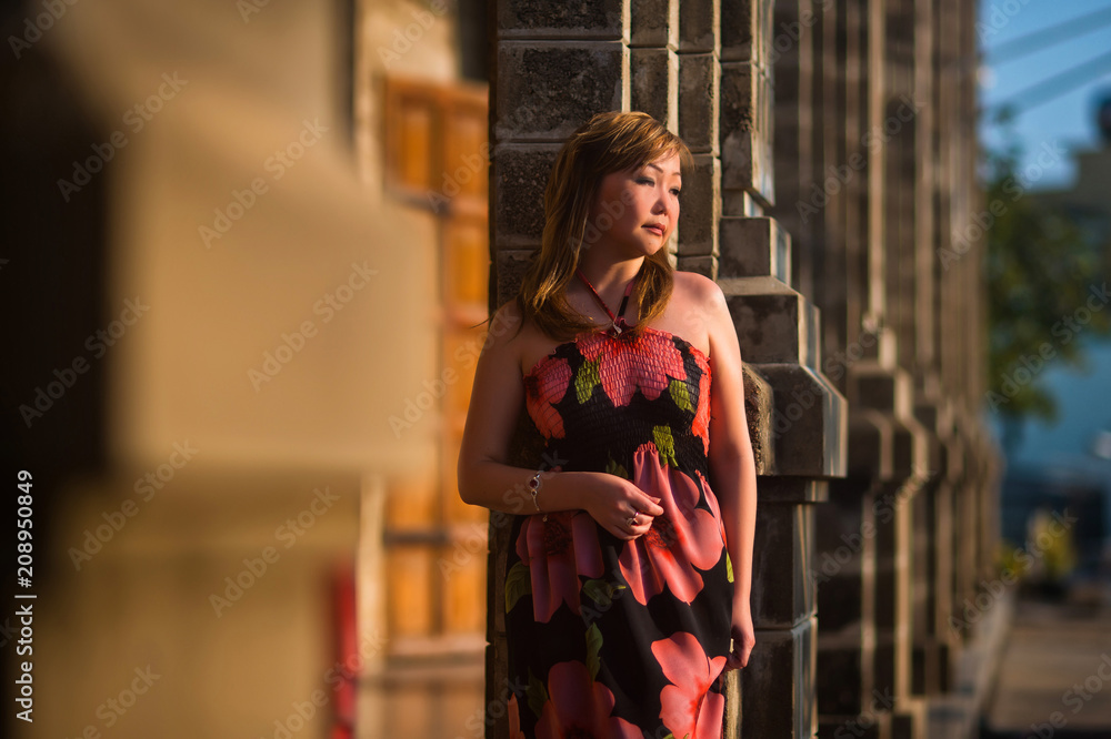 Portrait of an adult Asian woman, who in an elegant dress posing near the column at sunset