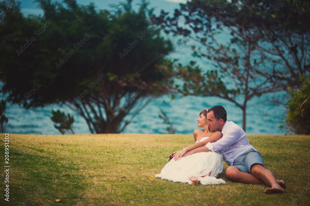Bride and groom hugging, sitting on the grass, against the beautiful tropical sea landscape