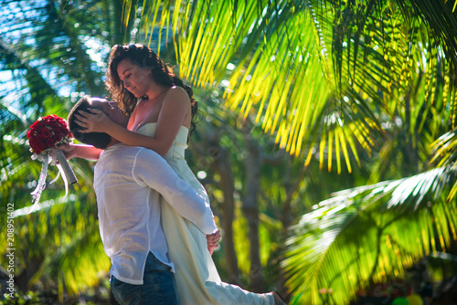 The groom holds the bride in his arms among the palm trees