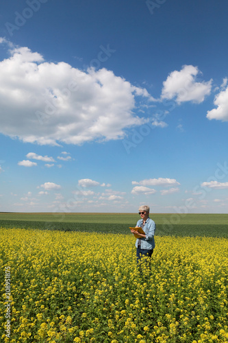 Female agronomist or farmer examining blossoming canola field and writing, rapeseed plant in early spring