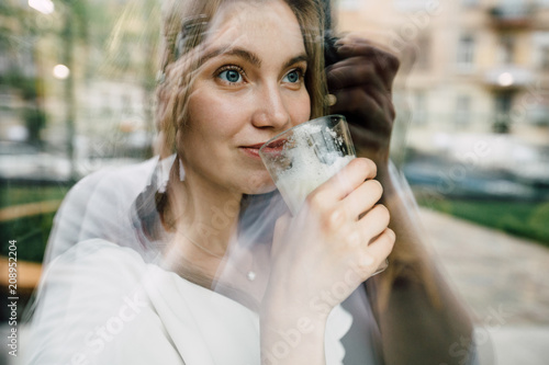 Young thoughtful blond woman drinking coffee looking through the window. Double exposure effect.