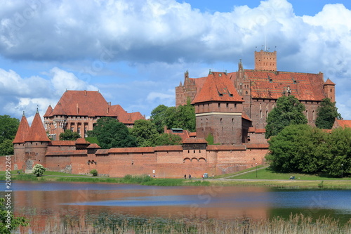 Fototapeta Naklejka Na Ścianę i Meble -  Zamek krzyżacki w Malborku, Polska, rzeka Nogat, zieleń wokół murów budowli, malownicze chmury na niebieskim niebie