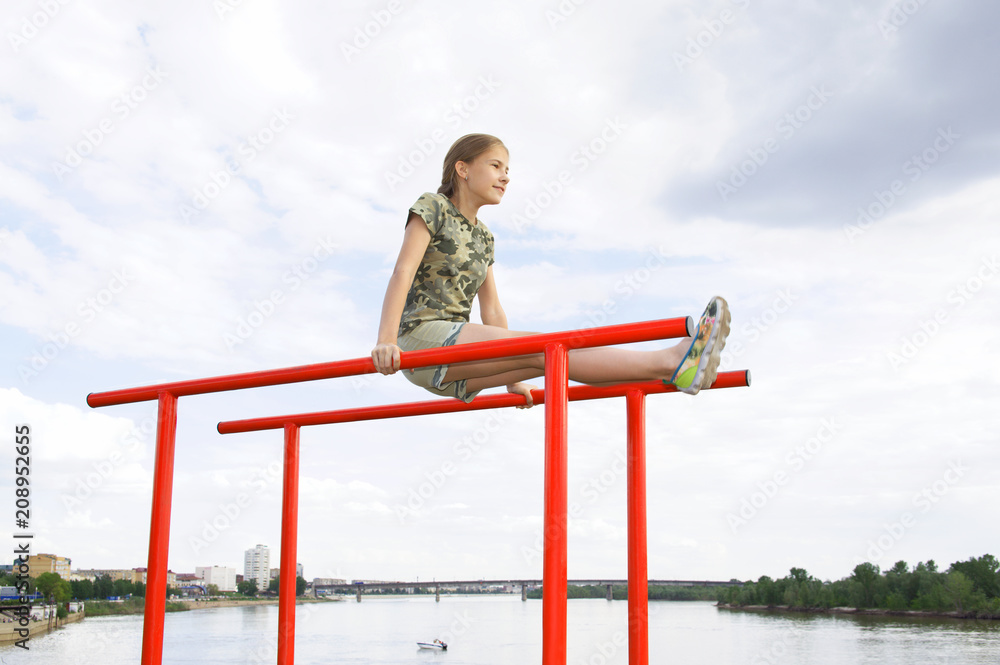 Smiling teenage girl doing gymnastic exercises on uneven bars in city conditions outdoors