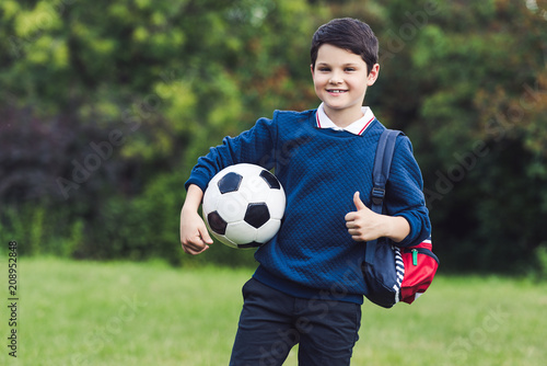 happy kid with soccer ball and backpack showing thumb up on grass field © LIGHTFIELD STUDIOS