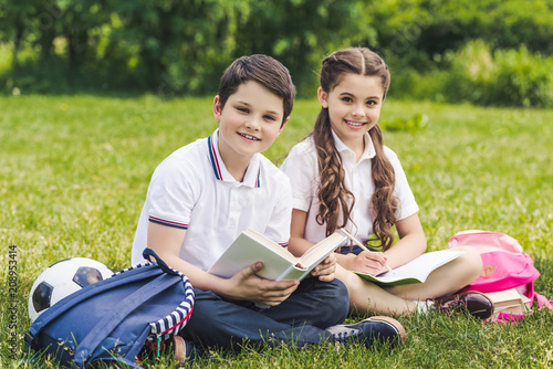 smiling schoolchildren doing homework together while sitting on grass in park and looking at camera