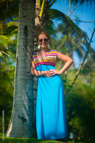 Beautiful fashionable young woman in dress and sunglasses standing near palm trees and smiling