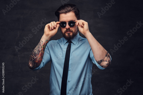 Portrait of a tattooed handsome middle-aged man with beard and hairstyle dressed in a blue shirt and tie, pose with hand on sunglasses.