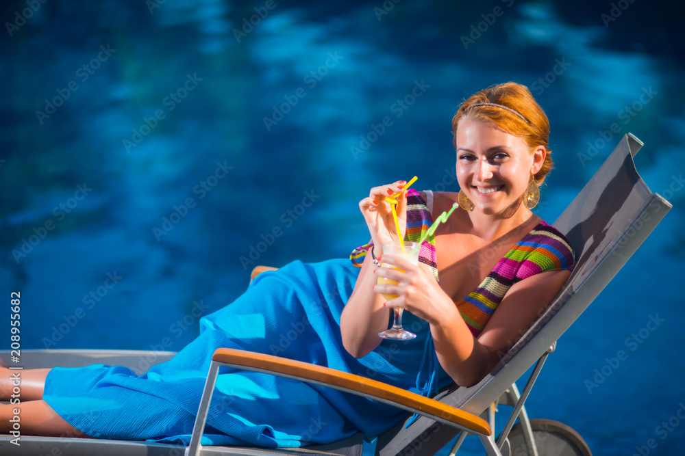Portrait of a young woman in a blue dress, who lies on a sun lounger by the pool against the palm trees, holding juice and smiling
