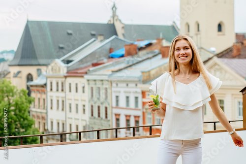 Young woman looking at camera, enjoying great view standing on the terrace of restaurant with soft drink glass..Travel, tourism and resting concept.