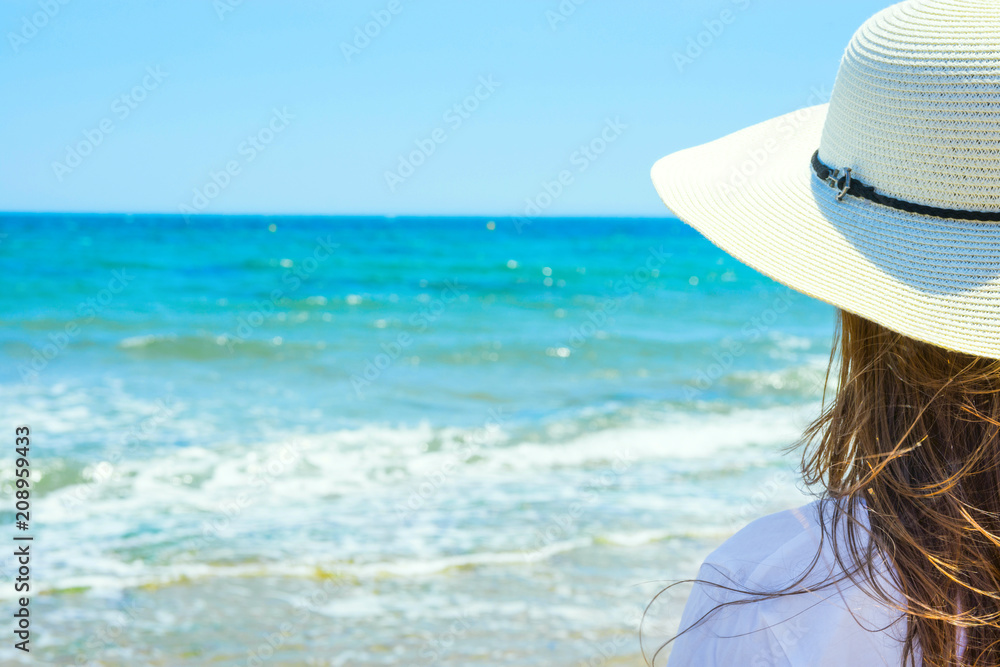 Young Attractive Caucasian Woman with Long Chestnut Hair in Hat Stands with Back On Sand Beach Looks at Turquoise Sea Horizon on Blue Sky Background. Tranquility Contemplation Summer Vacation