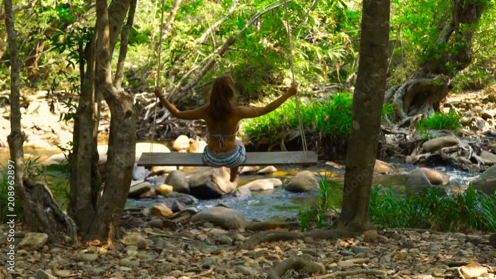 Woman on swing in rainforest on river shore back view. Young woman swinging on swing in jungle forest. Stony river in mountain background. Woman on swing in rainforest on river shore back view. Young woman swinging on swing in jungle forest. Stony river in mountain background.