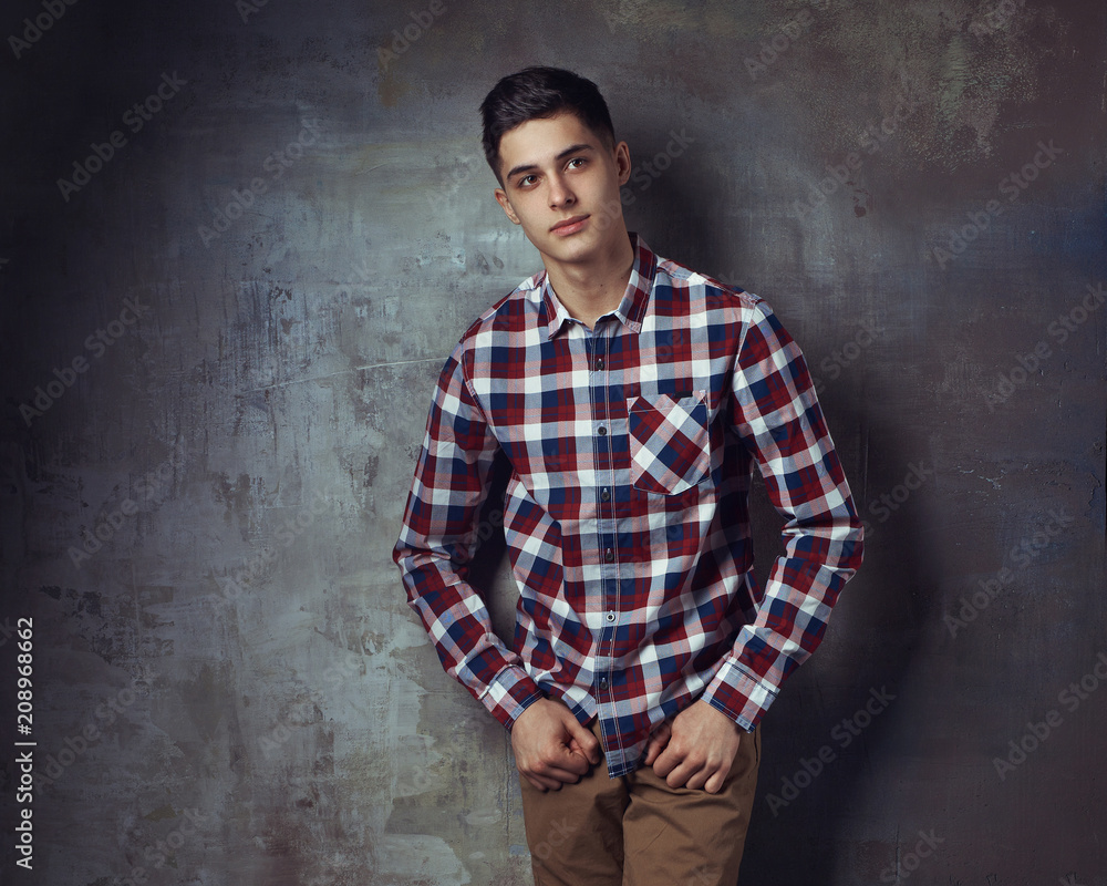 Portrait of young trendy handsome man with short dark hair wearing checkered shirt and brown trousers standing and posing against gray concrete wall