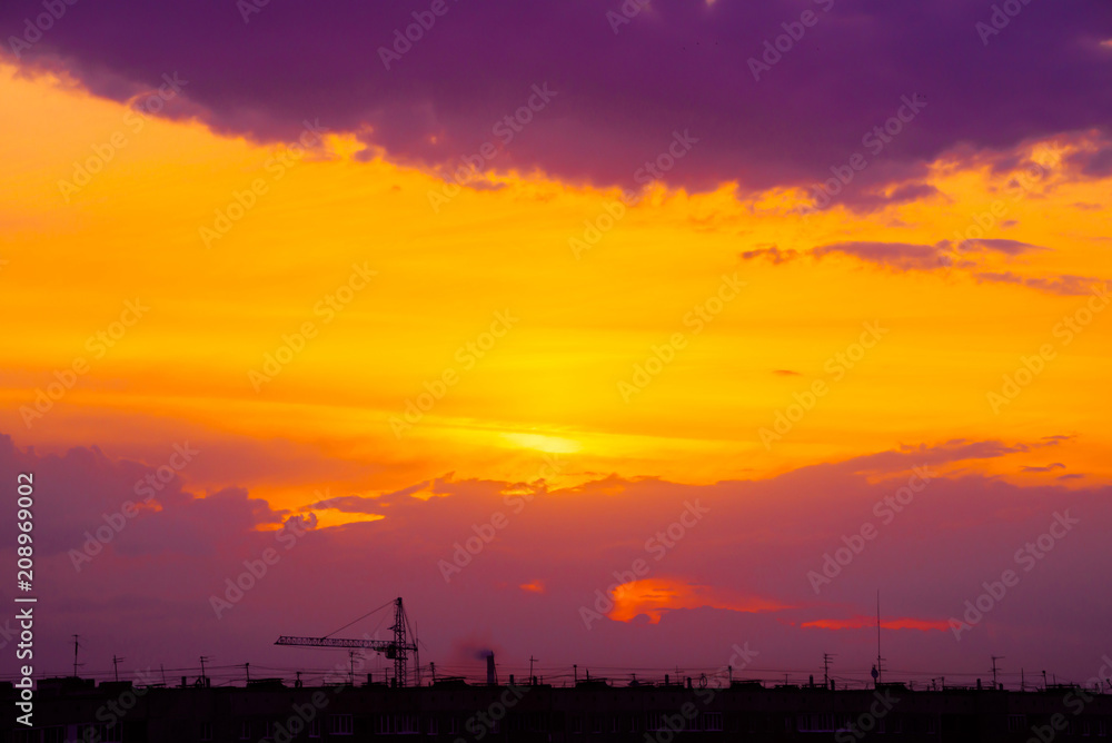 Magenta sunrise above silhouette of city buildings with tower cranes. Unusual cityscape with beautiful dawn. Picturesque warm city landscape with bright sunny sky. Orange and purple cloudy layer sky.