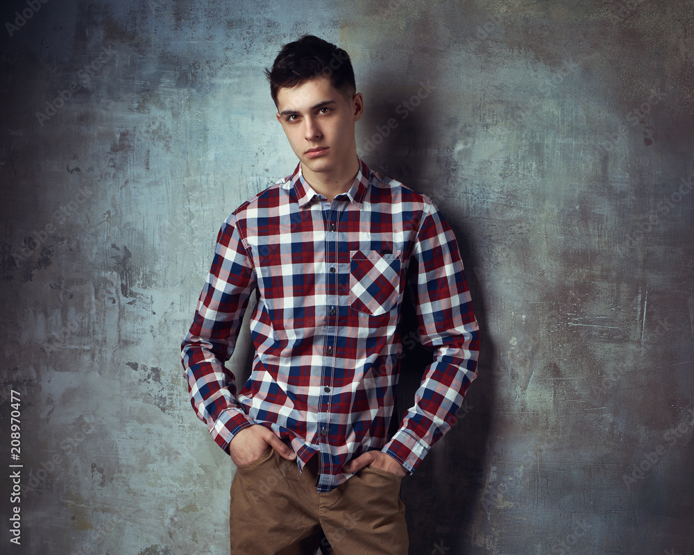 Portrait of young trendy handsome man with short dark hair wearing checkered shirt and brown trousers standing and posing against gray concrete wall