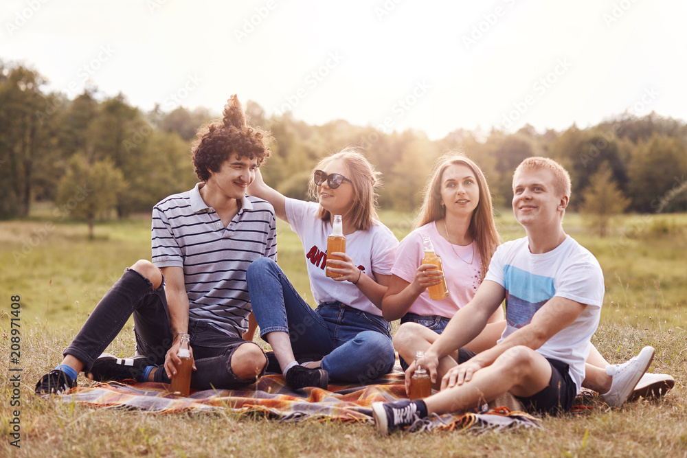 Fototapeta premium Outdoor shot of happy best friends have picnic in countryside, have fun, look into distance, sit on plaid on nature, show good relationship. Four female and male groupmates recreat outside together
