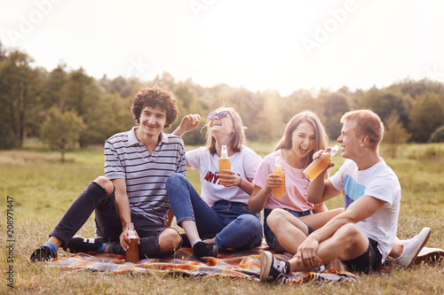Shot of happy teenagers laugh joyfully while have picnic outdoor, drink cold energy drinks, sit crossed legs, have fun, breath fresh air, pose outside, spend weekend on nature. Young and leisure © sementsova321