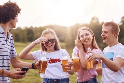 Summer time and leisure concept. Happy girls laugh at boy`s funny jokes, drink cold beverages, have cheerful expressions, spend spare time on summer field. Companionship and relationship concept