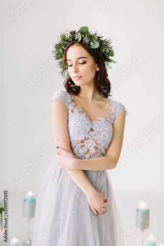 Beautiful young girl in a gray evening dress and a wreath of pine and eucalyptus on her head poses on the background of wedding scenery. Bridesmade poses in the studio before wedding ceremony. photo