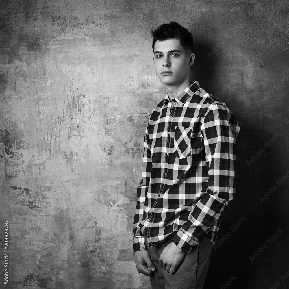 Obraz premium Portrait of young trendy handsome man with short dark hair wearing checkered shirt and brown trousers standing and posing against gray concrete wall