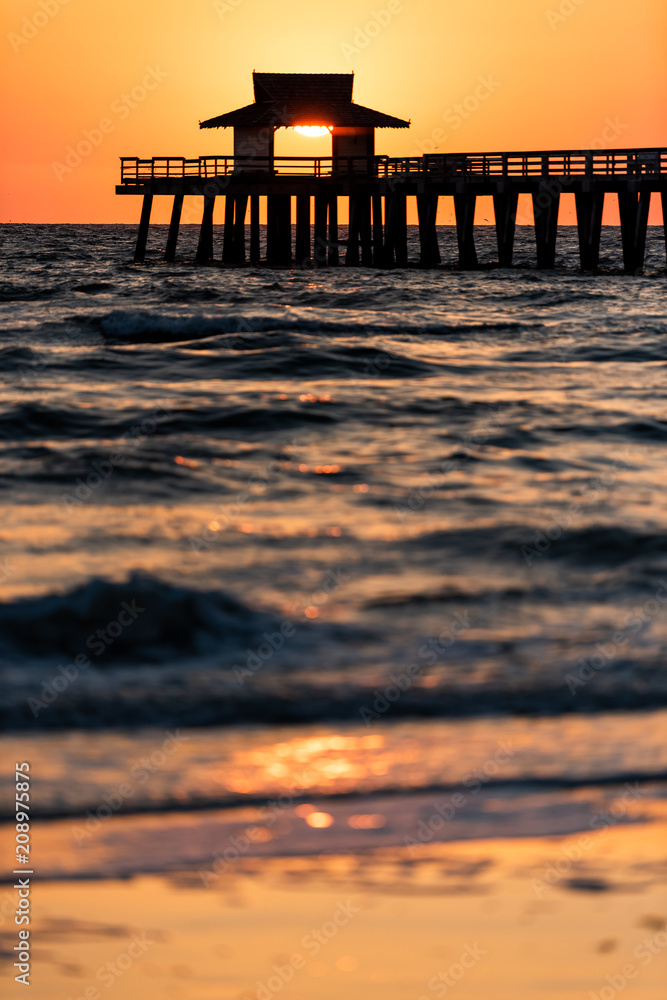 Naples, Florida yellow and orange sunset vertical view in gulf of Mexico with sun setting inside Pier wooden jetty, horizon and dark blue ocean waves Naples, Florida yellow and orange sunset vertical view in gulf of Mexico with sun setting inside Pier wooden jetty, horizon and dark blue ocean waves