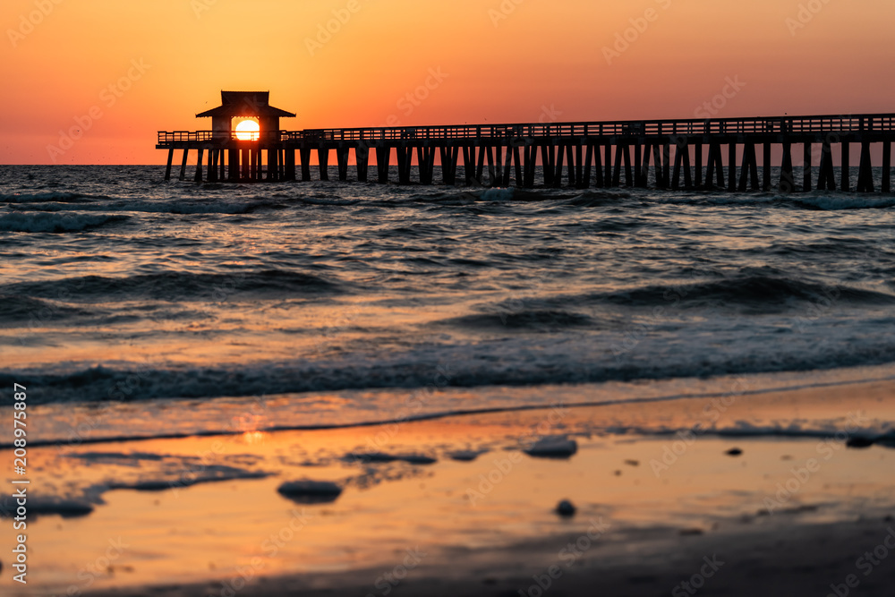 Naples, Florida red tide and orange sunset in gulf of Mexico with sun setting inside Pier, framing by wooden jetty, with birds flying over horizon and dark blue ocean waves