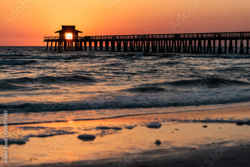 Naples  Florida red tide and orange sunset in gulf of Mexico with sun setting inside Pier  framing by wooden jetty  with birds flying over horizon and dark blue ocean waves