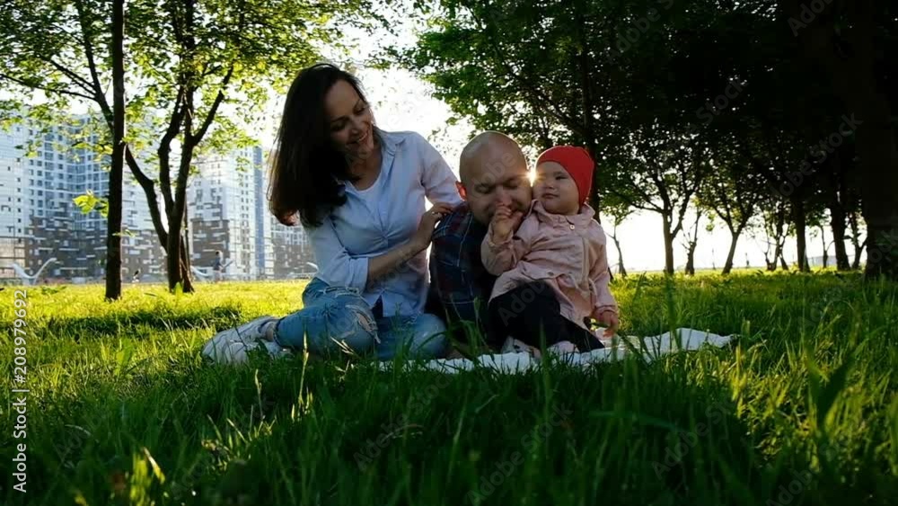 Happy family mother and father playing with a young child in summer park at sunset. Little baby girl sitting on grass laughing with parents. Happy family mother and father playing with a young child in summer park at sunset. Little baby girl sitting on grass laughing with parents.