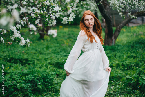Beautiful red-haired girl in a white dress among blossoming apple-trees in the garden.
