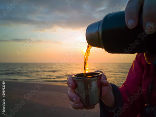 Pouring hot tea into thermos cup, sunset on background, nothern sea, adult womans hands.