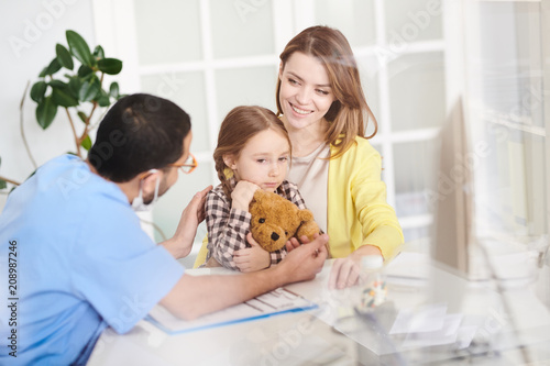 Back view portrait of young doctor talking to scared little girl hugging teddy bear sitting in mothers lap during consultation in modern clinic