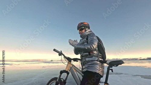 Woman is stay in bicycle on the ice. The girl is dressed in a silvery down jacket, cycling backpack and helmet. Woman is heating her hands with friction. Ice of the frozen Lake Baikal. The traveler is photo