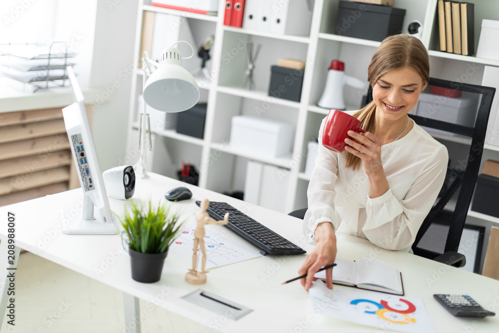 A young girl is sitting at a table in the office, holding a red cup in her hand and taking a sheet with a chart from the table. A young girl is sitting at a table in the office, holding a red cup in her hand and taking a sheet with a chart from the table.
