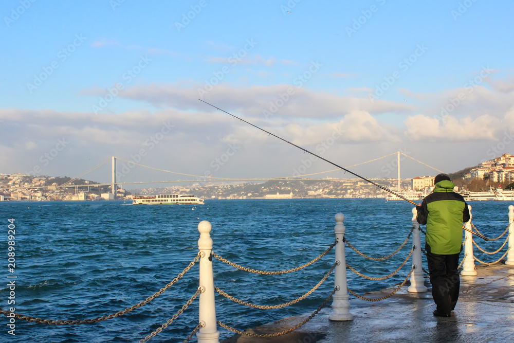 The man who wears green coat fishing in Istanbul in the bridge view.