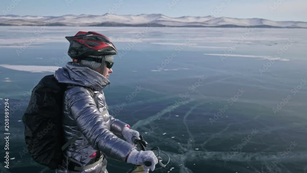 Woman is walking beside bicycle on the ice. The girl is dressed in a silvery down jacket, backpack and helmet. Ice of the frozen Lake Baikal. The tires on the bicycle are covered with special spikes Woman is walking beside bicycle on the ice. The girl is dressed in a silvery down jacket, backpack and helmet. Ice of the frozen Lake Baikal. The tires on the bicycle are covered with special spikes