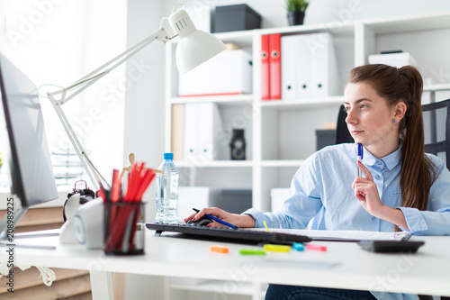 A young girl in the office sits at a table, holds a pen in her hand and works at the computer. photo