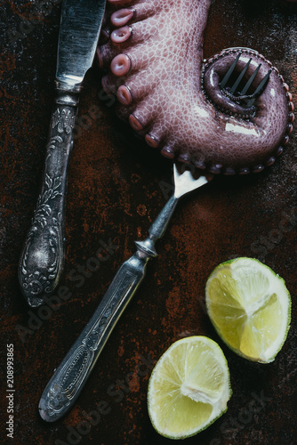 top view of big octopus tentacle with fork, knife and limes on rusty metal surface