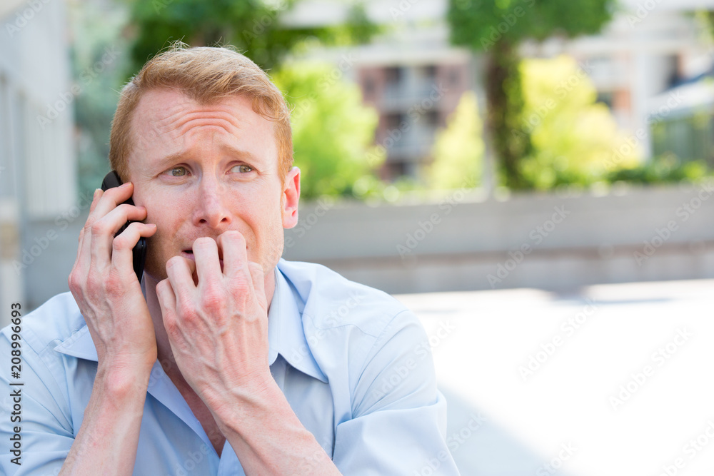 Closeup portrait, worried young man in blue shirt talking on phone to someone, looking gloomy, isolated outdoors outside background Closeup portrait, worried young man in blue shirt talking on phone to someone, looking gloomy, isolated outdoors outside background