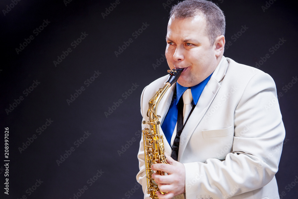 Fototapeta premium Music Concepts. Expressive Mature Caucasian Male Saxophone Player Posing in White Suite With Sax. Against Black Background.